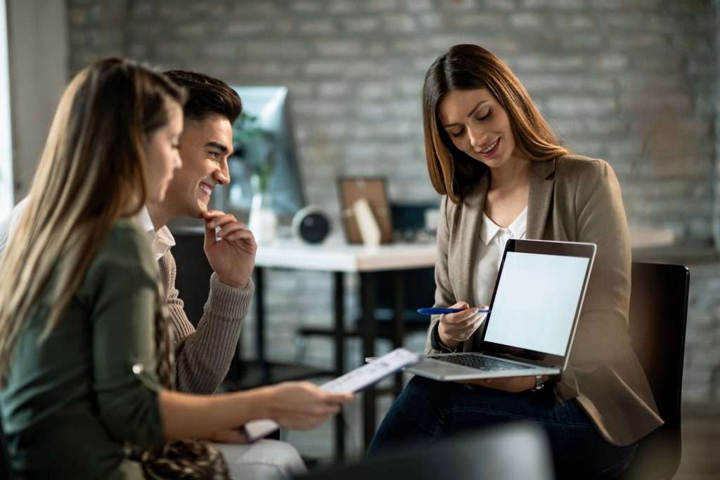 Confident young woman explain some details of document and pointing it with smile while sitting together with young couple at the desk in office